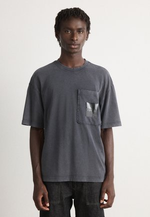 Dark gray cotton t-shirt with a loose fit, featuring a rectangular pocket with a glossy black finish and visible stitching details.