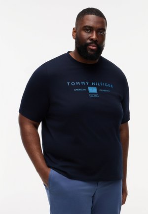 Skægget mand iført en sort Tommy Hilfiger T-shirt med blå tekst og blå bukser, stående med den ene hånd i lommen mod en hvid baggrund.