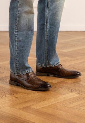 Persoon die bruine, gestructureerde leren nette schoenen draagt met donkerblauwe jeans, staand op een lichte houten parketvloer.