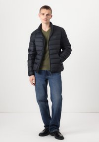 Schwarze Steppjacke mit Reißverschluss vorne, getragen über einem grünen V-Ausschnitt-Shirt. Kombiniert mit blauen Jeans und schwarzen Schuhen, stehend vor einem schlichten Hintergrund.