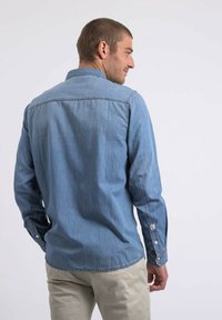 Chemise en denim bleu clair à manches longues, avec des poignets à bouton et un col classique. Présente un détail de couture minimal aux épaules et à l'ourlet.