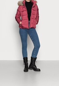 Czerwona puchowa kurtka z kapturem wyłożonym futrem, kieszeniami na zamek i pikowanym wzorem, zestawiona z dżinsami i czarnymi militarnymi butami.