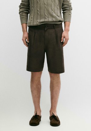 Mann trägt dunkelbraune maßgeschneiderte Shorts, olivgrünen Zopfpullover und braune Mokassin-Schuhe, steht vor einem schlichten hellen Hintergrund.