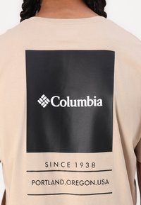 Beige Baumwoll-T-Shirt mit einem großen schwarzen quadratischen Grafikdesign, auf dem "Columbia" steht, sowie einem Text darunter. Es hat eine glatte Textur und ein einfaches Design.
