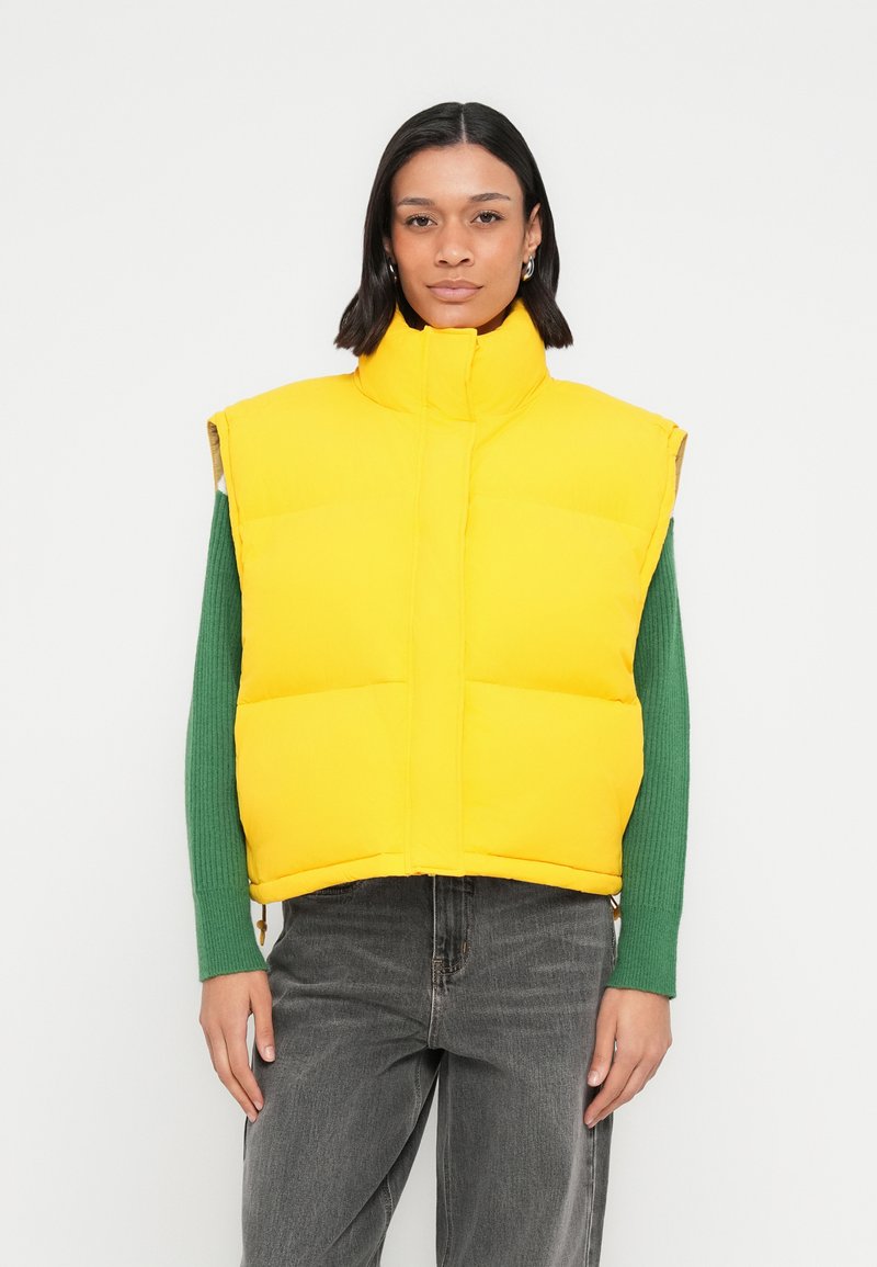Bright yellow puffer vest with a high collar, zippered front, and sleeveless design, paired with a green ribbed long-sleeve shirt underneath.
