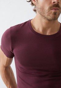 Burgundi rövid ujjú póló, testhez simuló fazonnal és kerek nyakkal. Simaság jellemzi, egyszínű, és minimális varrások észlelhetők.