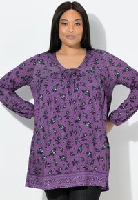 Fioletowa bluzka w kwiatowy wzór, z długimi rękawami, dekoltem w kształcie litery V oraz marszczeniem z przodu. Dół bluzki ozdobiony dekoracyjną lamówką.