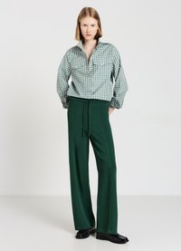 Frau mit blonden Bob-Haarschnitt, die ein grün karriertes Button-Up-Hemd und dunkelgrüne weite Hosen trägt, steht vor einem weißen Hintergrund.