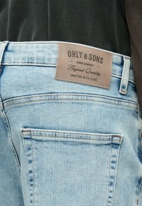 Šviesiai mėlyni džinsai pagaminti iš džinso audinio, turi odinę etiketę su užrašu „ONLY & SONS“ ir siūlių akcentus. Matomos galinės kišenės su klasikiniu dizainu.