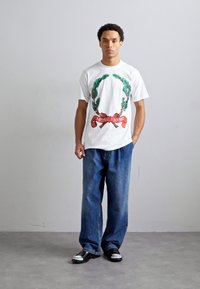 White graphic t-shirt featuring green laurel leaves and a red ribbon with "Moschino" text, paired with loose blue denim jeans and metallic shoes.