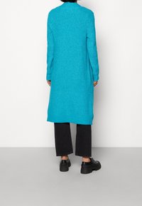 Lungo cardigan turchese a maglia con un design testurizzato, dotato di colletto e polsini a costine, abbinato a pantaloni neri e scarpe nere spesse.