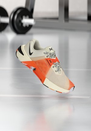 Chaussure de sport beige clair et orange vif avec logo Swoosh noir, lacets à motifs, et poids de gym flous en arrière-plan.