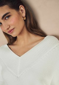 Vit ribbad tröja med V-ringning, med en texturerad tyg. Modellen bär guldiga droppformade örhängen.