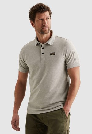 Mann mit kurzen braunen Haaren, der ein hellgraues Polohemd mit kurzen Ärmeln und olivgrüne Hose trägt, steht mit einer Hand in der Tasche und blickt nach links.