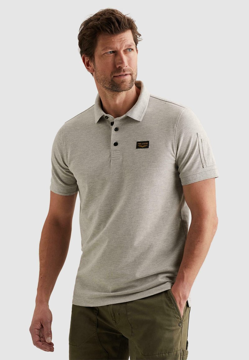 Muškarac s kratkom smeđom kosom, obučen u svijetlosivu polo majicu kratkih rukava i maslinasto zelene hlače, stoji s jednom rukom u džepu i gleda lijevo.