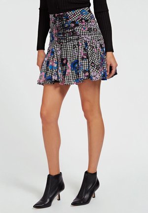 Kobieta ubrana w krótką spódnicę z kwiatowym i pepitkowym wzorem oraz sznurowanym przodem, w połączeniu z czarnym prążkowanym topem z długim rękawem i czarnymi botkami na obcasie.