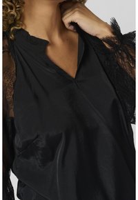Femme portant un chemisier en satin noir avec des manches en dentelle, tenant le col près de son cou, montrant partiellement des cheveux bouclés.