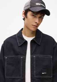 Noor mees, kes kannab musta samet mütsi, millel on logo "Karl Lagerfeld Jeans", ja musta jakki valge õmblusega ning esikottidega, valge särgiga.