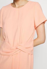 Robe de couleur pêche en tissu léger, avec un col rond, des manches courtes et un détail noué à la taille.