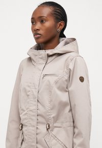 Veste beige clair avec fermeture éclair sur le devant, taille ajustable par cordon de serrage, poches latérales et capuche. Patch de logo sur l'épaule gauche. Texture du tissu lisse.