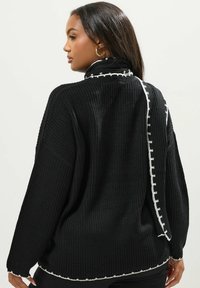 Musta neulepaita, jossa on rento malli, korkea kaulus ja kontrastiväriset valkoiset ompeleet selkäpuolella ja helmassa.