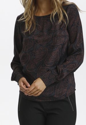 Kobieta ubrana w ciemną bluzkę z wzorem paisley na długich rękawach z marszczonymi mankietami oraz czarne spodnie z widocznymi kieszeniami na zamek.
