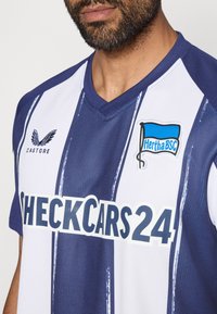 Sinine ja valge triibuline jalgpallipaita V-kaelusega, millel on "CHECKCARS24" logo ja Hertha BSC embleem. Valmistatud tekstureeritud kangast.