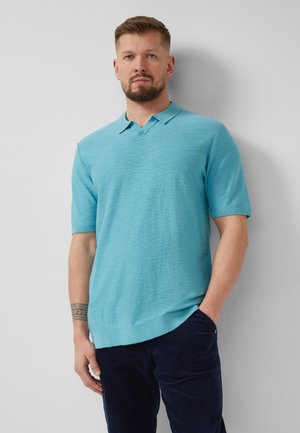 Mann mit Bart und kurzen Haaren, der ein hellblaues kurzärmeliges Poloshirt und dunkle Hosen trägt, steht vor einer hellgrauen Wand.