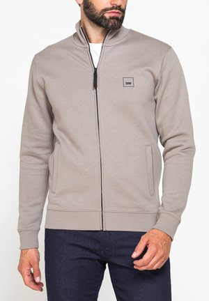 Uomo con giacca beige con zip sopra camicia bianca e pantaloni scuri, in piedi davanti a uno sfondo chiaro e semplice.