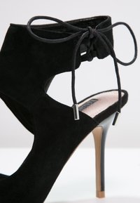 Chaussure à talon haut en daim noir avec côtés ouverts, fermeture à lacets à la cheville et embouts métalliques sur les lacets.