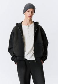 Sweat à capuche noir zippé avec une coupe décontractée, grandes poches avant, porté sur une chemise henley blanche, associé à un pantalon noir et un bonnet gris.