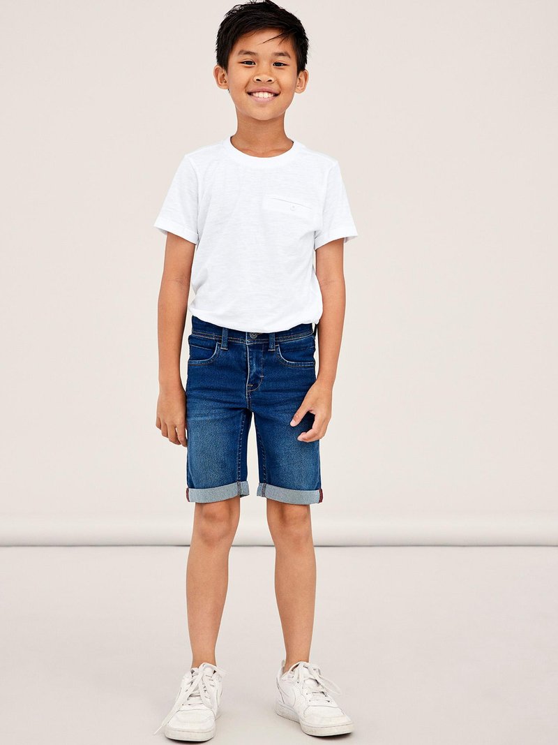 White short-sleeve t-shirt with a pocket, paired with rolled-cuff blue denim shorts. White sneakers complete the outfit.