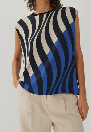 Mouwloos topje met golvende zwarte, beige en blauwe strepen, gedragen met lichtbeige broek met hoge taille. Persoon staat met één hand in de zak.