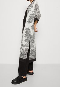 Long black and white kimono-style jacket with paisley print, three-quarter sleeves, and side slits, paired with black trousers and slip-on shoes.