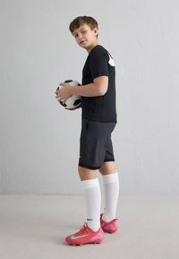 Pois mustas spordirõivastuses ja valgete põlvesokkidega, hoides jalgpalli käes, seljas roosad jalgpalliketsid, seisab lihtsa helehalli seina ääres.