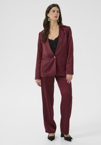 Donna che indossa un completo in satin bordeaux con giacca a un bottone su un top in pizzo nero, abbinato a décolleté nere a punta.