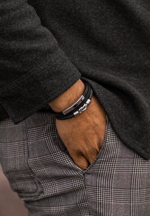 Black braided leather bracelet with a silver clasp, worn over a gray long-sleeve shirt, with plaid patterned pants visible.
