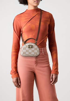 Sac à main tan avec un motif de logo, en forme arrondie, et un fermoir doré. Sangle brun réglable. Le modèle porte un haut orange à manches longues et un pantalon rose.