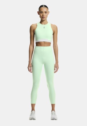 Kvinde stående iført en lysegrøn atletisk crop top og leggings med høj talje samt hvide sneakers, på hvid baggrund.