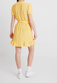 Robe florale jaune avec des manches bouffantes courtes, une taille cintrée et un ourlet volanté, assortie à des baskets blanches et des chaussettes de cheville.