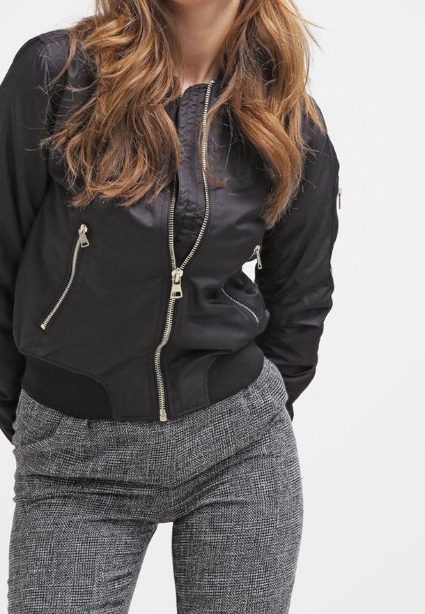 Femme aux longs cheveux portant une veste noire zippée et un pantalon gris à motifs, debout devant un fond blanc uni.