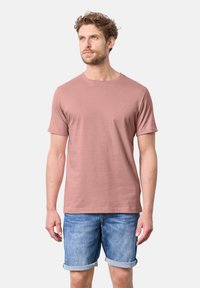 T-shirt da uomo a maniche corte in rosa polveroso, realizzato in tessuto morbido con colletto rotondo e un piccolo logo sul petto. Indossato con pantaloni corti di denim.