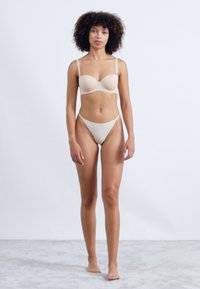 Soutien-gorge rembourré beige avec des fines bretelles, associé à un string taille basse assorti. Texture lisse, design minimaliste sur un fond clair.