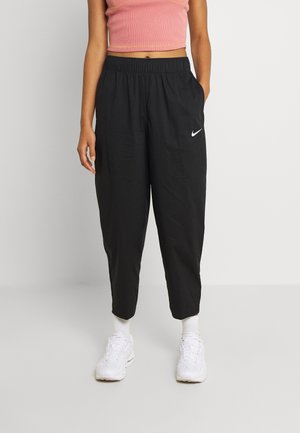 Vrouw draagt zwarte Nike joggingbroek, roze kort tanktopje, witte sokken en witte sneakers, staat met één hand in de zak.