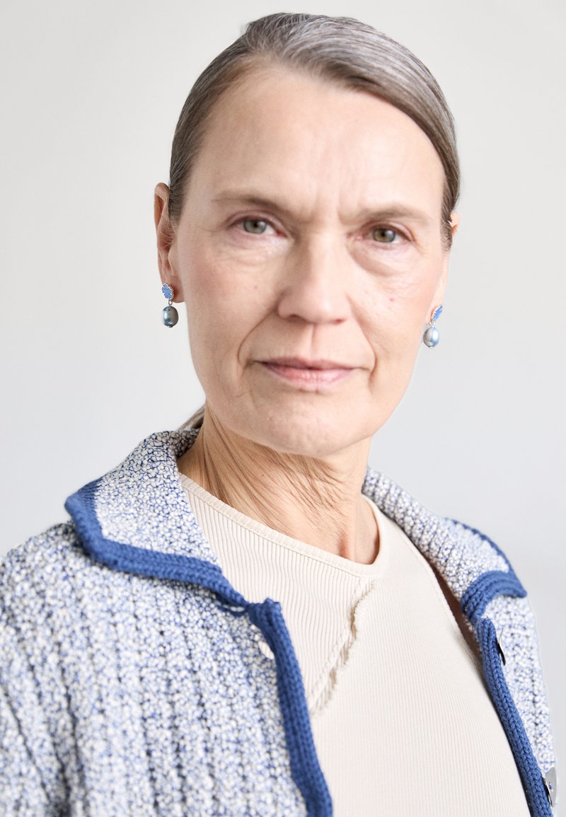 Vaaleansininen teksturoitu takki, jossa on kauluksen malli ja siniset yksityiskohdat. Beige ribbitoppi takin alla. Ripustettavat korvikset, joissa on sinisiä yksityiskohtia.