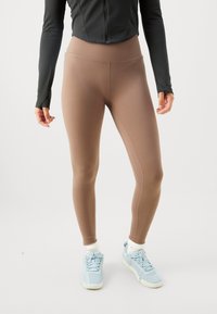 Kvinna klädd i beige högmidjade leggings, vita strumpor, ljusblå sneakers och en svart långärmad zip-up-tröja som står på en enkel bakgrund.