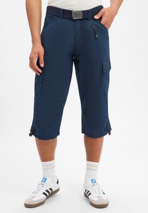 Man wearing navy blue cargo shorts with multiple pockets, white socks, and white Adidas sneakers with black stripes.