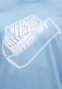 Ανοιχτό μπλε ύφασμα με λευκό στιλιζαρισμένο λογότυπο Nike που περιλαμβάνει γράμματα σε πλάτος και ένα swoosh με τρισδιάστατο εφέ.