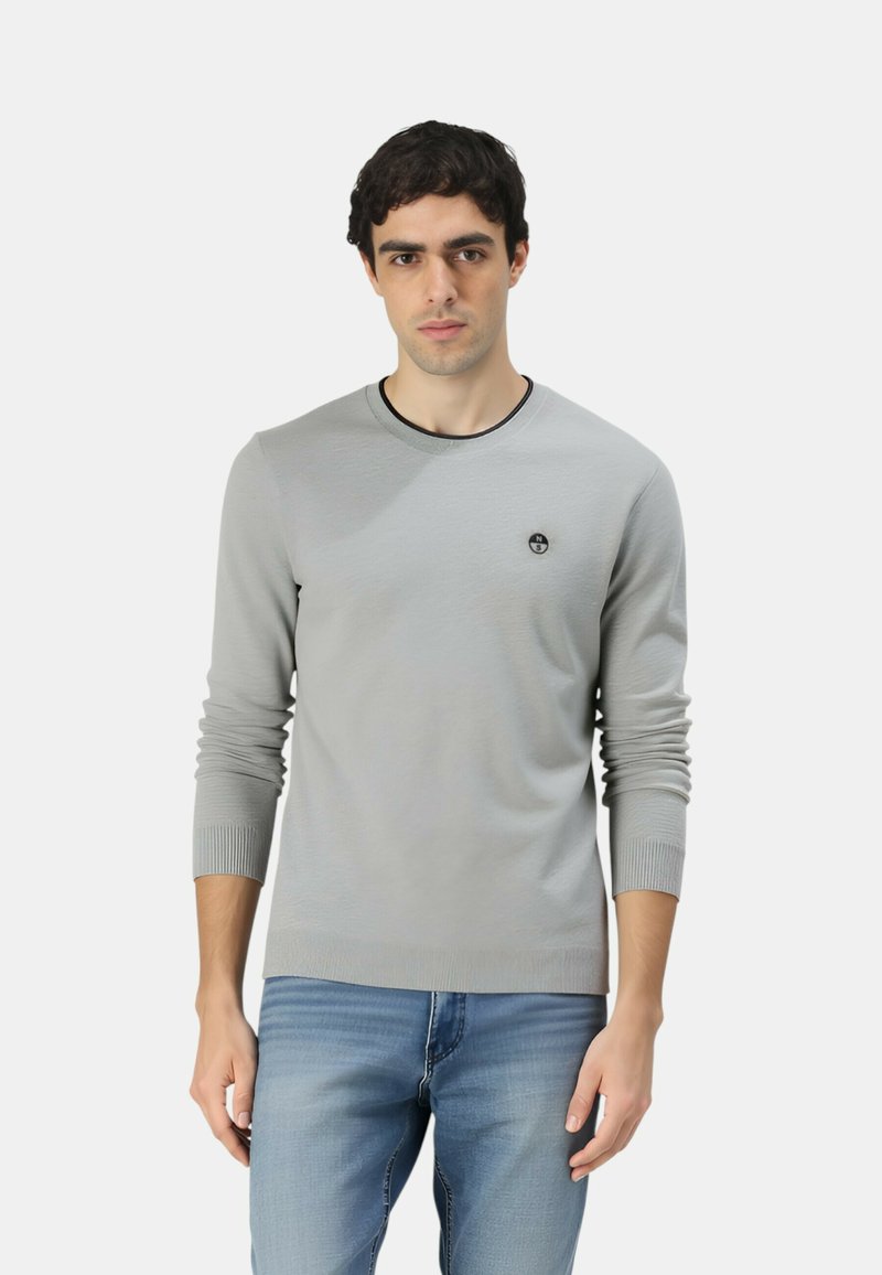 Maglione grigio lavorato a maglia con maniche lunghe, collo tondo, decorato da un piccolo logo sul petto, abbinato a jeans azzurri chiari.