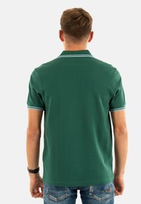 Camisa polo verde, de mangas cortas, con cuello acanalado y ribete azul claro, vista trasera, combinada con jeans azul desgastados.